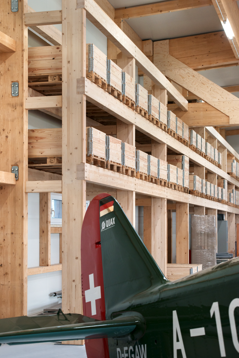 Innenansicht des Hangars mit mehrstöckigem Holzregalsystem an der Seitenwand und dem Heck eines grünen Kleinflugzeugs im Vordergrund.