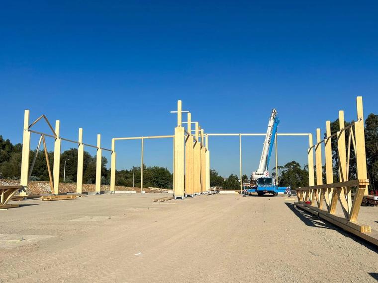 Weite Baustellenansicht mit aufgestellten Holzstützen, Mobilkran und am Boden lagernden Fachwerkbindern