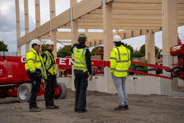 Vier Monteure des Teams von LIGNA systems in Warnwesten im Gespräch vor dem entstehenden Holztragwerk