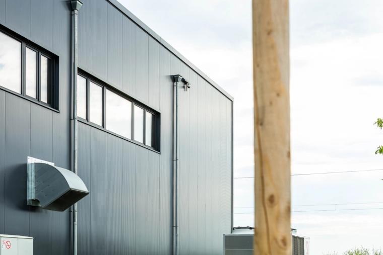 Seitliche Außenfassade der Halle aus grauen Profiltafeln mit Fensterband, im Vordergrund ein Holzpfosten und eine Lüftungshaube