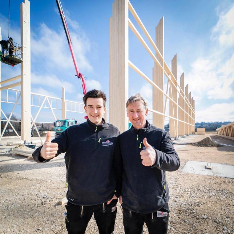 Zwei Mitarbeiter in Berufskleidung auf der Baustelle in Krauchenwies, im Hintergrund die bereits aufgerichteten Brettschichtholz-Stützen des Hallenrohbaus.