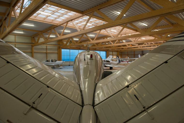 Close-up of a light aircraft tail section with propeller in the foreground, with further aircraft and the hangar's timber load-bearing structure behind.