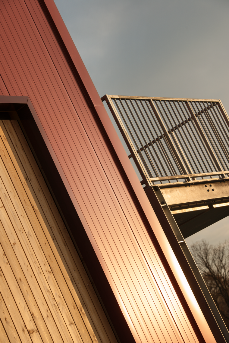 Fassadendetail des Hangars mit senkrechter Holzverkleidung, rotem Metallband darüber und dem Geländer der außenliegenden Stahltreppe im Gegenlicht.