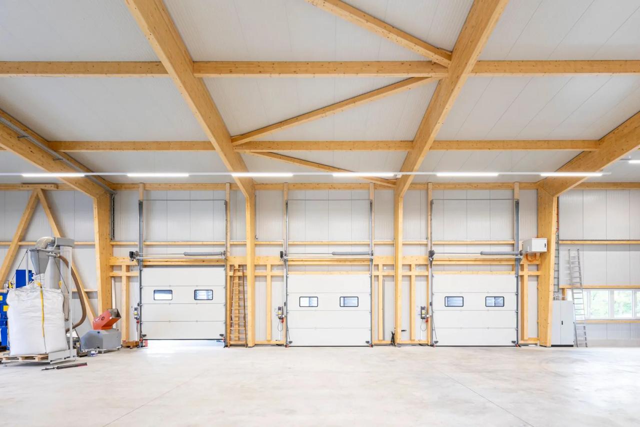 Binnenaanzicht van een systeemhal met houten kolommen en wijd overspannende houten liggers, drie sectionaaldeuren in de kopgevel