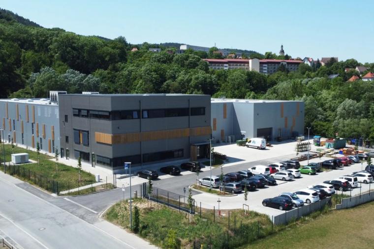 Luftaufnahme des fertiggestellten Produktionskomplexes mit zweigeschossigem Bürotrakt, Holzlattenelementen an der Hallenfassade und angrenzendem Parkplatz im Grünen.