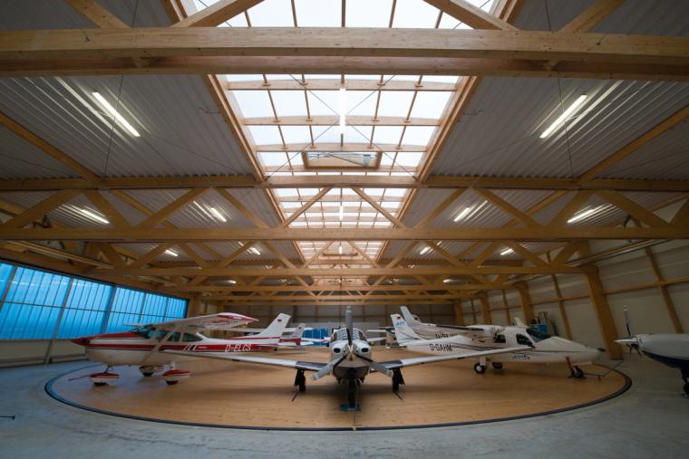 Wide-angle interior of the hangar with light aircraft parked on a circular floor marking beneath the exposed timber truss and ridge skylight.