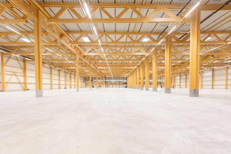 Symmetrische Innenansicht der Logistikhalle entlang der Längsachse mit beidseitigen Holzstützenreihen, Fachwerkdach und Blick bis zur Stirnwand.