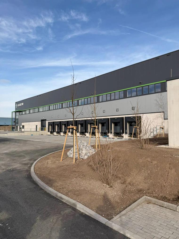 Außenansicht der Logistikhalle mit mehreren Ladetoren und frisch gepflanzten Bäumen auf dem Vorplatz.
