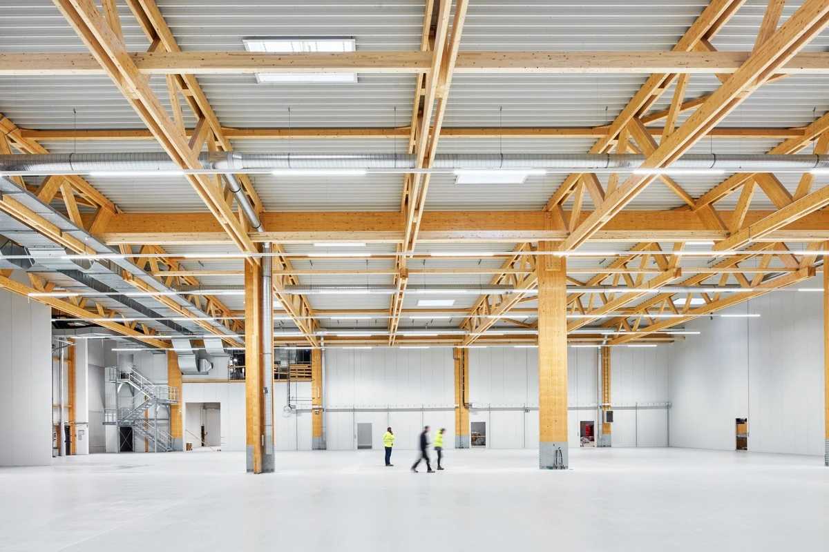Innenansicht der Produktionshalle mit freistehendem Holzbinder-Tragwerk auf massiven Holzstützen, hellem Bodenbelag und drei Personen in Warnwesten im Vordergrund.