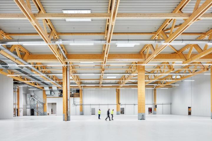 Innenansicht der Produktionshalle mit freistehendem Holzbinder-Tragwerk auf massiven Holzstützen, hellem Bodenbelag und drei Personen in Warnwesten im Vordergrund.