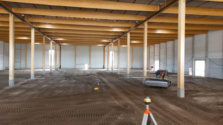Innenraum der Halle unter dem geschlossenen Dach, Holzstützen und Träger im Raster, Vermessungsgerät und Raupe auf dem Planum