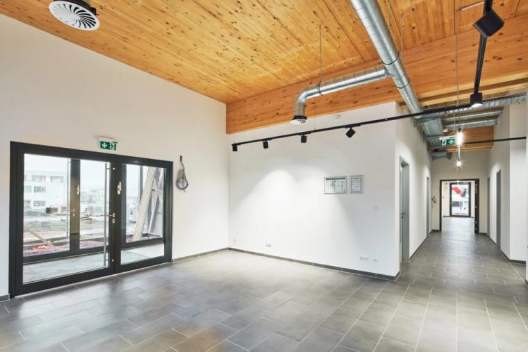 Empfangs- und Erschließungsbereich im Bürotrakt mit sichtbarer Holzdecke, freiliegender Lüftungsleitung, Schienenbeleuchtung und Glasdoppeltür mit Blick zur Baustelle.