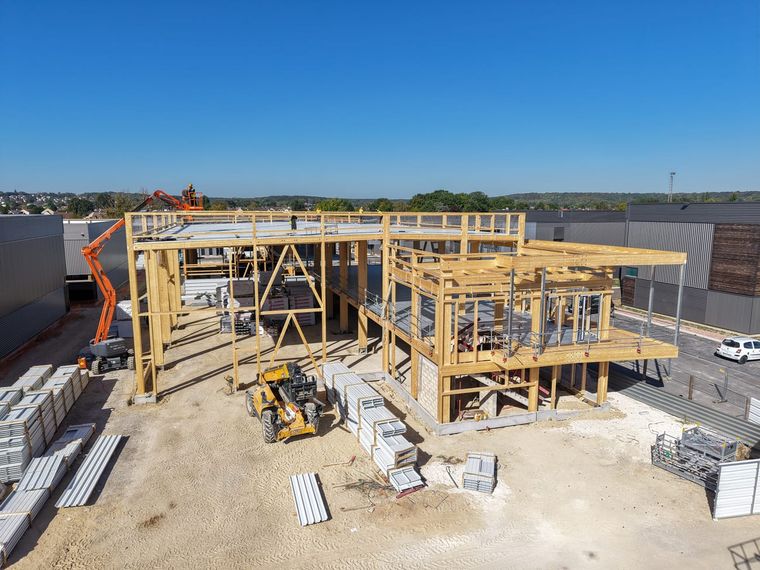 Vue aérienne par drone du chantier de Courtry : gros œuvre en angle à ossature bois avec poteaux visibles et planchers en porte-à-faux, une nacelle en action, à côté des bâtiments voisins achevés à la façade métallique sombre