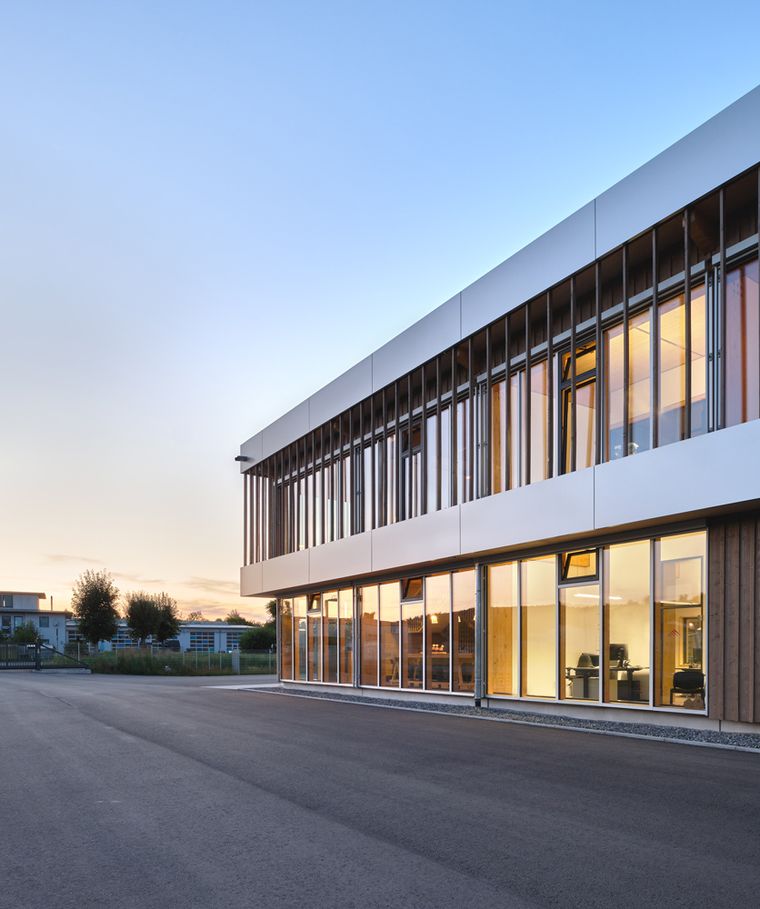 Seitenansicht des Gebäudes in der Abenddämmerung, erleuchtete Büroräume hinter der Glasfront und Holzfassadenverkleidung im warmen Licht.