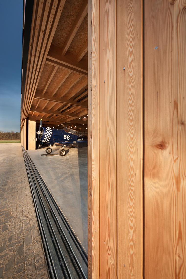 Blick durch ein geöffnetes Holzschiebetor in das Hangarinnere, wo ein blau-weißes Kleinflugzeug unter der Holzdecke steht.
