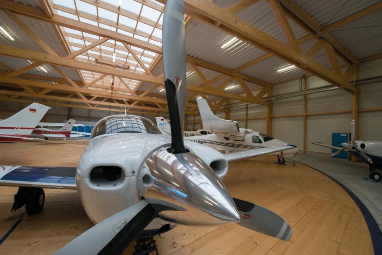 Close-up of a light aircraft nose and propeller, with the timber columns and a roof skylight of the hangar visible to the side.