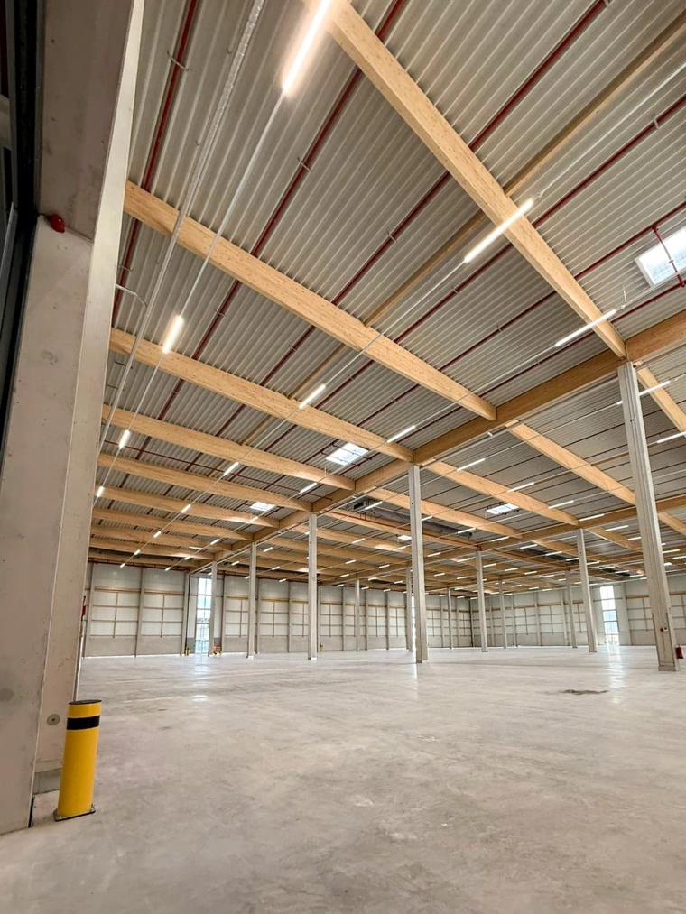 Weitwinkelaufnahme des leeren Hallenschiffs mit freiliegenden Holzbindern im Dachtragwerk und Betonboden.