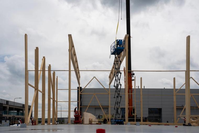 Aufrichtung des Holztragwerks: schlanke Brettschichtholz-Stützen und erste Binder stehen, eine blaue Scherenarbeitsbühne und ein Kran daneben.