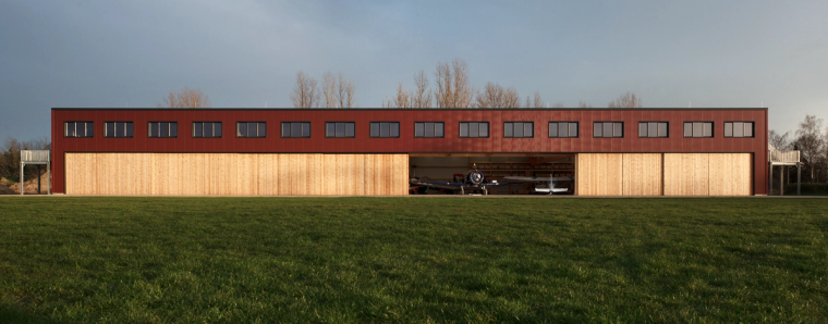 Lange Torfront des Flugzeughangars in Paulinenaue, ein Schiebetor steht weit offen; oben rote Fassade mit Fensterband, unten Holzverkleidung.