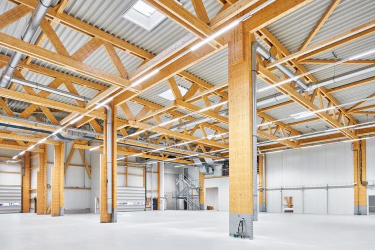 Weitwinklige Innenansicht der Produktionshalle mit sichtbarem Holzbinder-Dachtragwerk auf Holzstützen, Treppenaufgang zur Mezzanine-Ebene und geschlossenen Sektionaltoren.