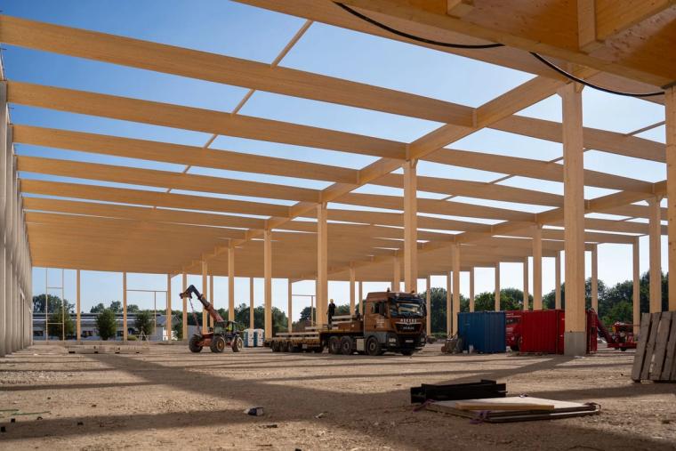 Blick in die Halle unter dem fertig montierten Tragwerk aus Holz vor blauem Himmel, ein Sattelzug liefert Bauteile auf die Bodenplatte