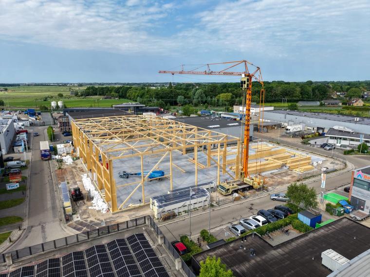 Luftaufnahme der Baustelle in Voorhout: das hölzerne Tragskelett der Produktionshalle mit Fachwerkbindern steht, daneben ein orangefarbener Turmdrehkran im Gewerbegebiet.