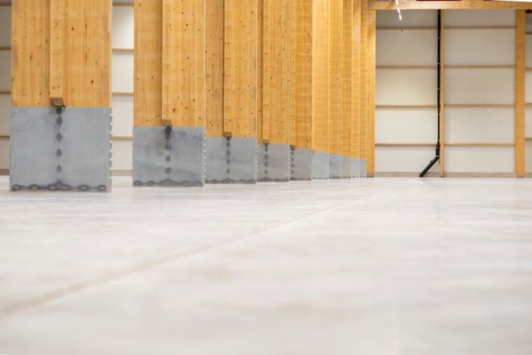 Bodennahe Innenaufnahme entlang einer Reihe von Holzstützen mit sichtbaren Betonpodesten an den Stützenfüßen und dem Betonboden im Vordergrund.