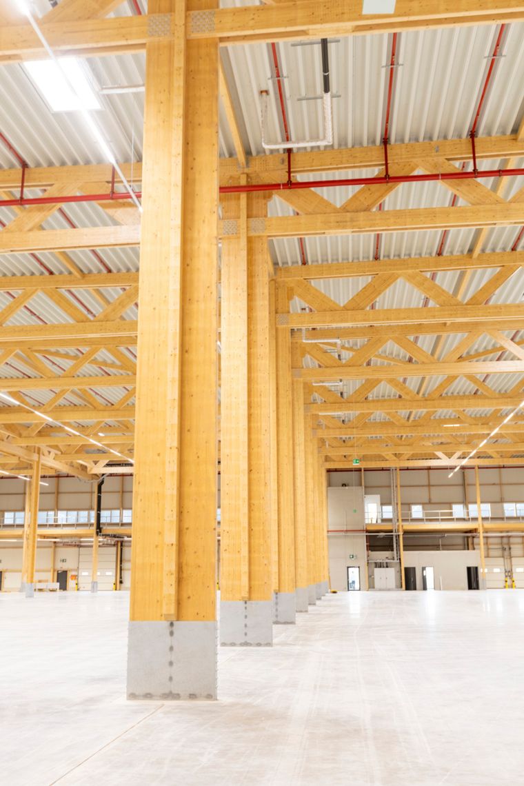 Hochformatige Innenaufnahme neben einer massiven Holzstütze mit Betonpodest, Blick entlang der Stützenreihe bis zum Ende der Halle.