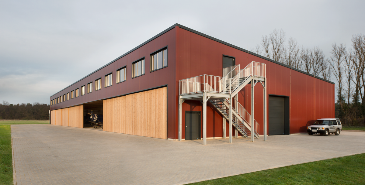 Flugzeughangar mit roter Metallfassade, naturbelassener Holzverkleidung im Erdgeschoss und außenliegender Stahltreppe auf befestigtem Vorplatz.