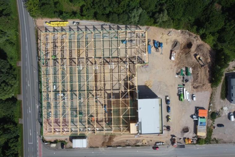 Drohnenaufnahme der Baustelle in der Rohbauphase: das Holztragwerk steht vollständig, die Dachfläche ist teilweise beplankt, auf dem Baufeld Baumaschinen und Materiallager.
