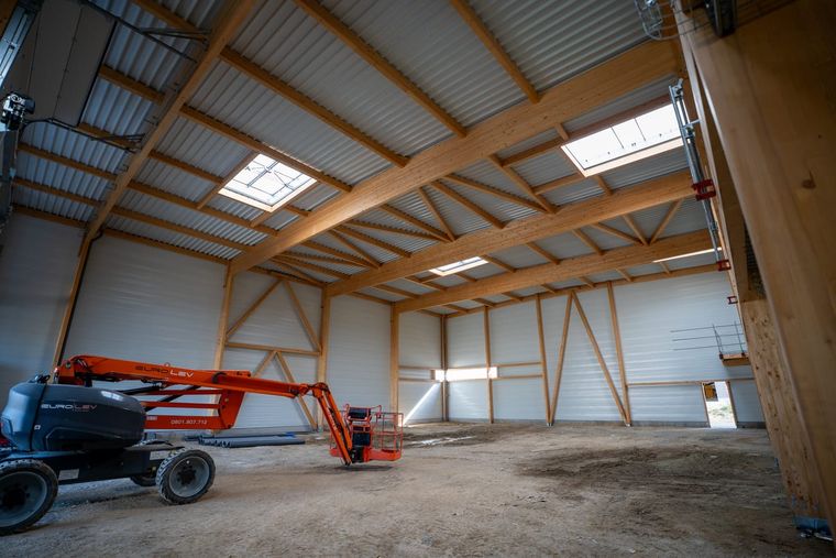 Vue de l’extérieur sous le débord de toiture d’un hall à Courtry : charpente ouverte avec pannes et contre-fiches en bois, la pose de la façade est en cours sur le long-pan