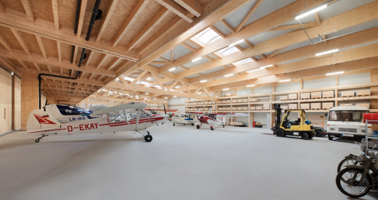 Helle Hangarinnenansicht mit sichtbaren Holzbindern, Dachoberlichten und mehreren geparkten Leichtflugzeugen, darunter ein rotes Modell im Vordergrund.