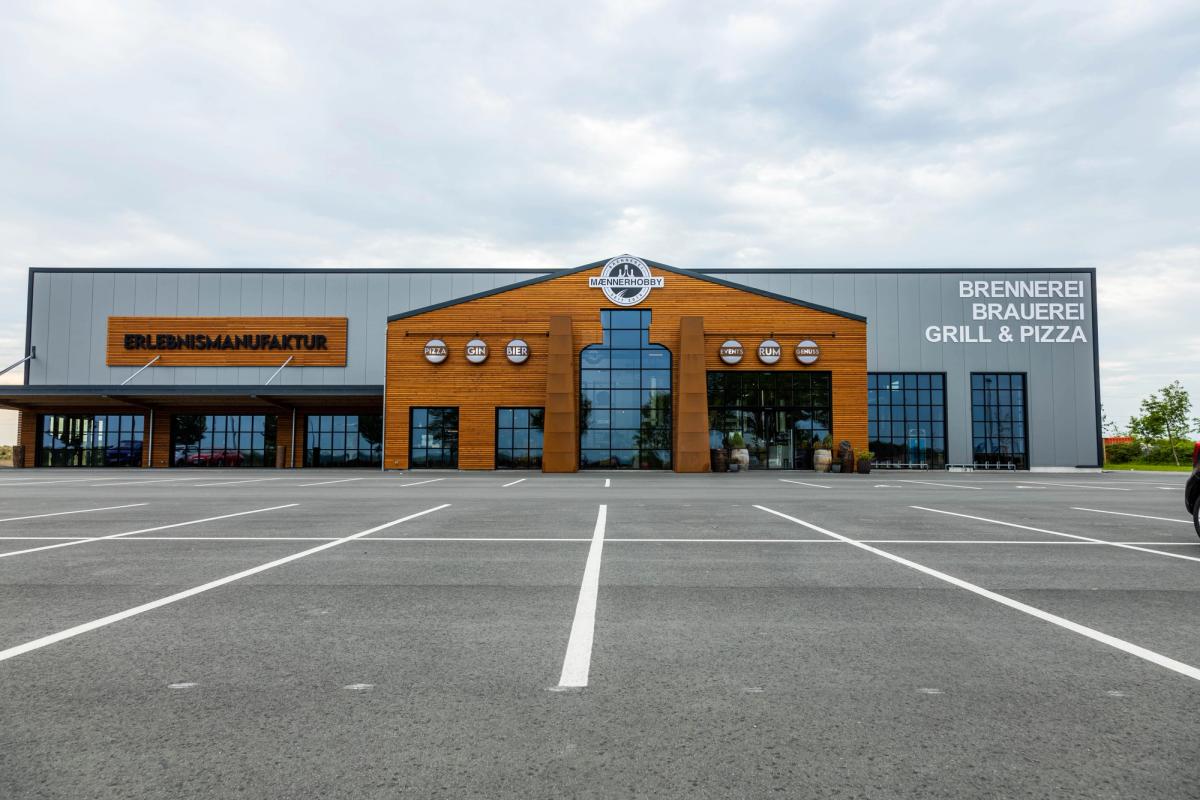 Frontansicht der Brauereihalle: langer grauer Baukörper mit holzverkleidetem Mittelgiebel, den Schriftzügen „Erlebnismanufaktur" und „Brennerei Brauerei Grill & Pizza" und großem Parkplatz davor