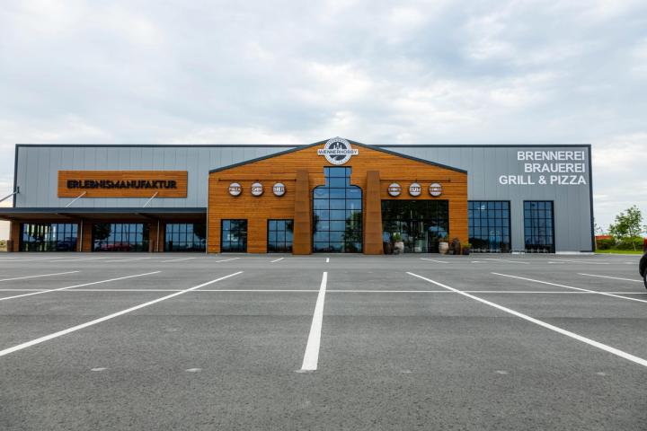 Frontansicht der Brauereihalle: langer grauer Baukörper mit holzverkleidetem Mittelgiebel, den Schriftzügen „Erlebnismanufaktur" und „Brennerei Brauerei Grill & Pizza" und großem Parkplatz davor