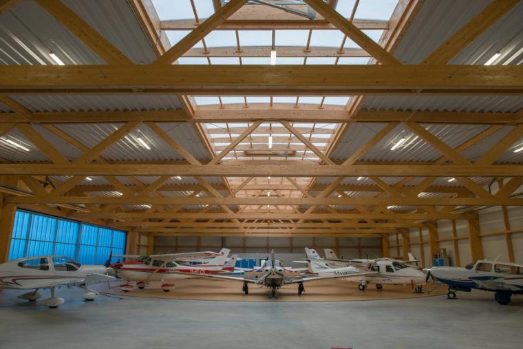 Front-on interior view of the hangar showing a row of parked light aircraft, a freestanding timber structure, and a ridge skylight band.