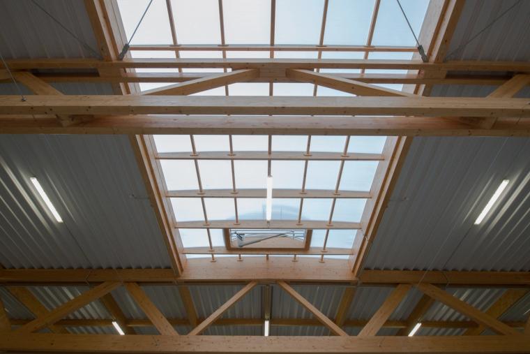 Detail shot of the roof structure from below: timber truss members with metal purlins and a central skylight of translucent panels.