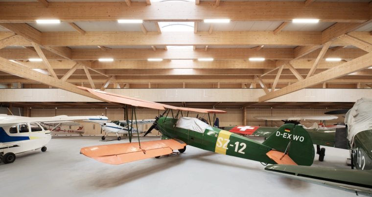 Hangarinneres mit einem grünen Doppeldecker-Oldtimer im Vordergrund und weiteren Kleinflugzeugen unter dem sichtbaren Holztragwerk.