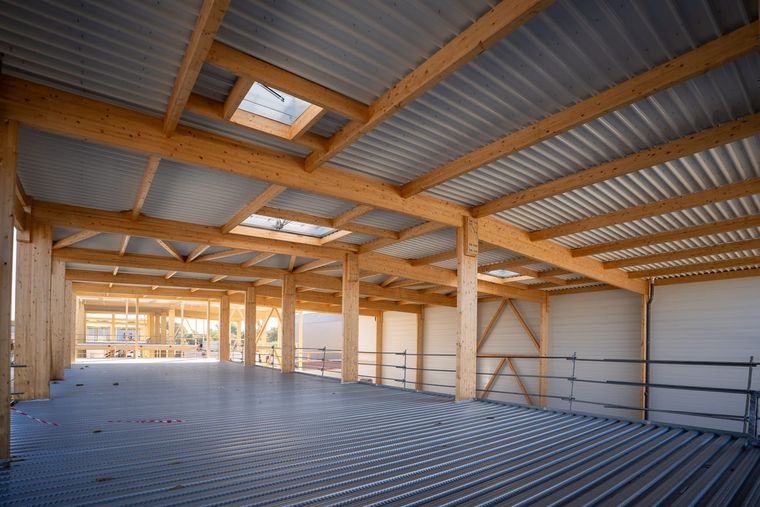 Vue intérieure à l’étage à Courtry : rangée de poteaux en bois clair avec poutres principales, plafond en bac acier, bandes de toiture translucides au-dessus, panneaux de façade partiellement montés sur le côté