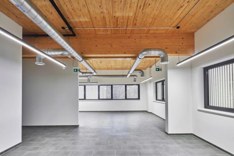 Heller Büroraum auf der Mezzanine-Ebene mit sichtbarer Holzdecke, freiliegenden Lüftungsrohren und Fenstern mit Blick zur Produktionshalle.