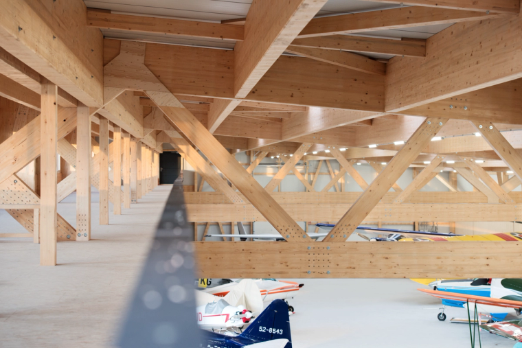 Innenansicht des Hangars mit sichtbarem Brettschichtholz-Fachwerkdach und mehreren abgestellten Kleinflugzeugen auf dem Hallenboden.