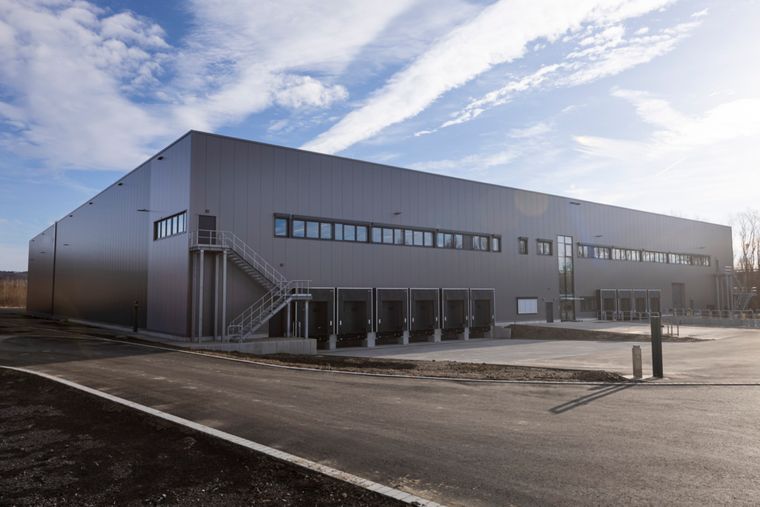 Außenansicht der Logistikhalle von der Zufahrtseite mit mehreren Laderampen, einem außenliegenden Treppenaufgang und grauer Metallfassade.