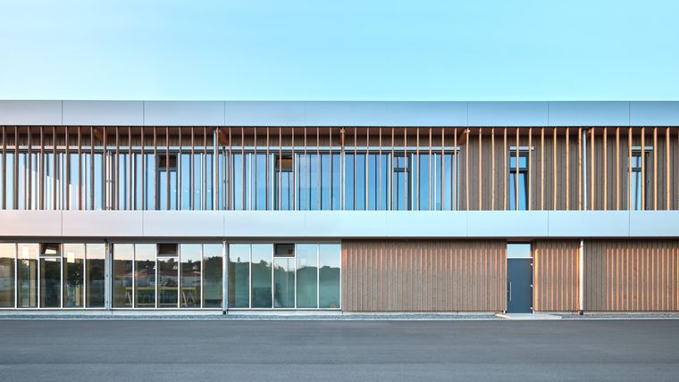 Frontansicht der zweigeschossigen Holz-Glas-Fassade mit vertikalen Holzlamellen und durchgehender Fensterfläche im Tageslicht.