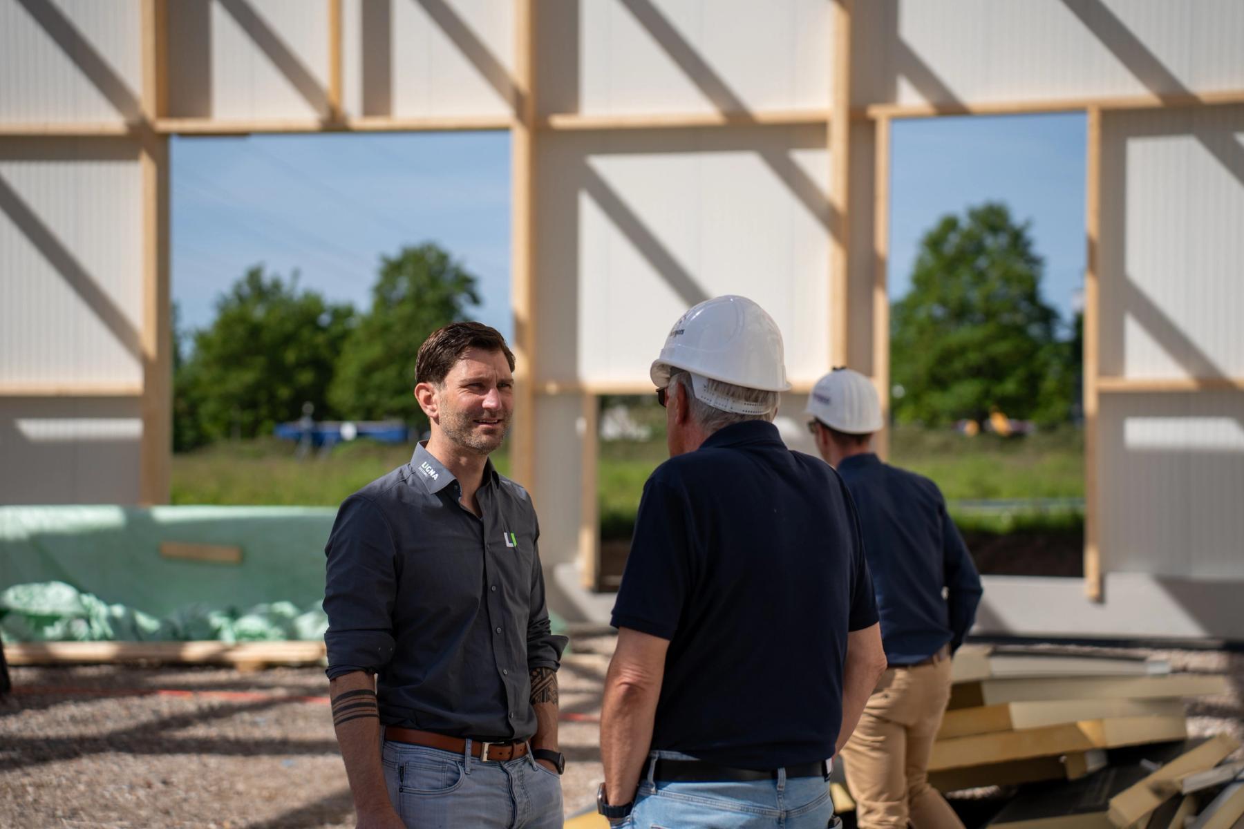 Deux hommes en discussion sur le chantier devant l’ossature en bois d’un hall en construction, l’un portant un casque.