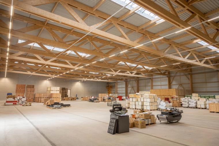 Innenansicht der Logistikhalle mit freiliegendem Holzbinder-Dachtragwerk und großzügiger Hallenfläche, auf der Waren auf Paletten zwischengelagert sind.