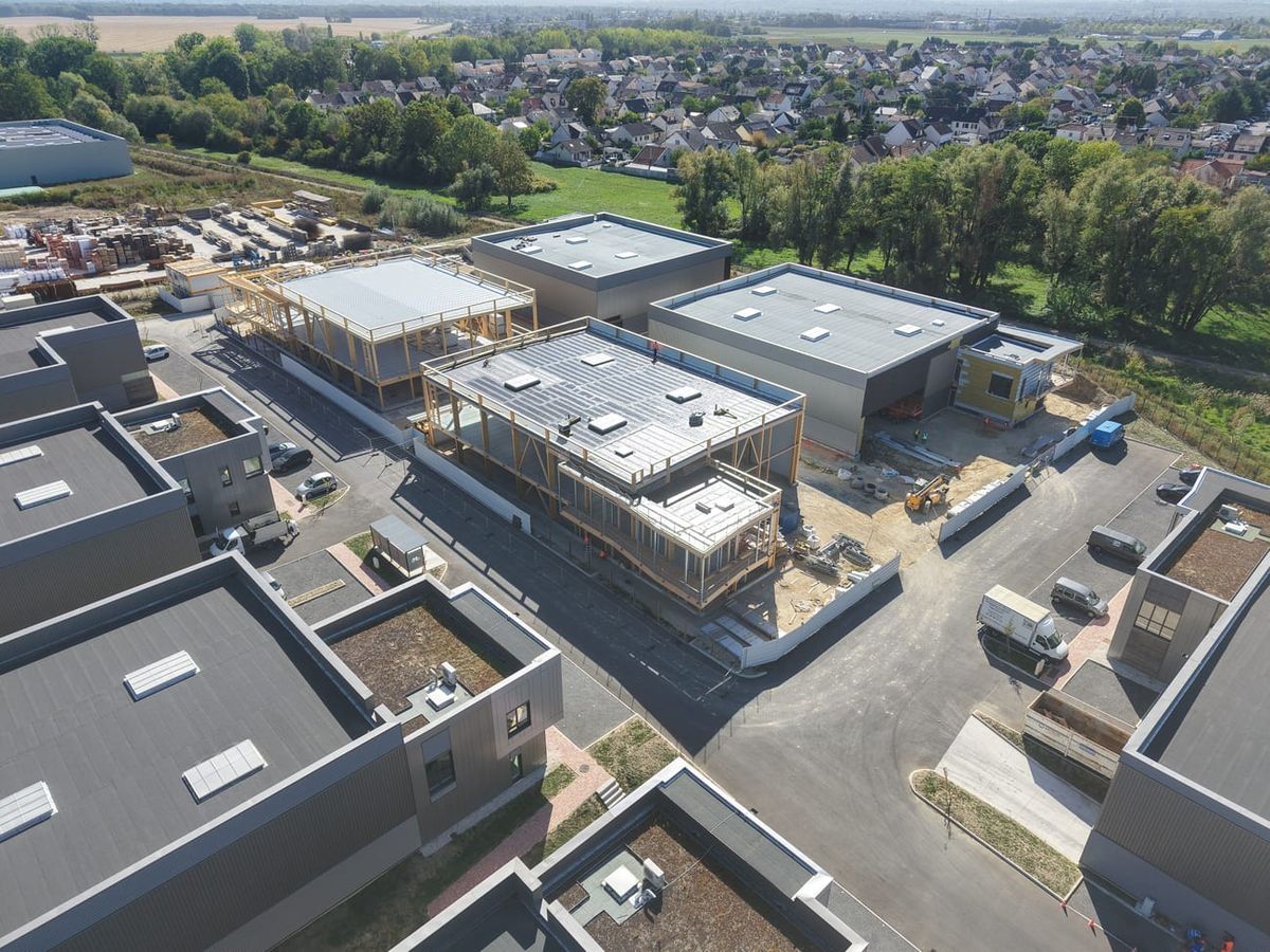 Drohnenaufnahme des Gewerbeparks in Courtry: der Rohbau der Gebäude in Holzbauweise mit heller Dachabdichtung im Zentrum, umgeben von fertiggestellten Nachbargebäuden, im Hintergrund Wohngebiet und Felder