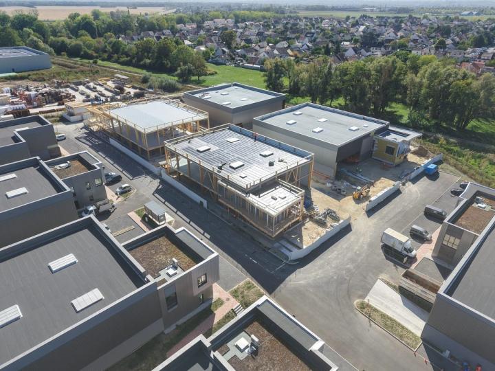 Drohnenaufnahme des Gewerbeparks in Courtry: der Rohbau der Gebäude in Holzbauweise mit heller Dachabdichtung im Zentrum, umgeben von fertiggestellten Nachbargebäuden, im Hintergrund Wohngebiet und Felder