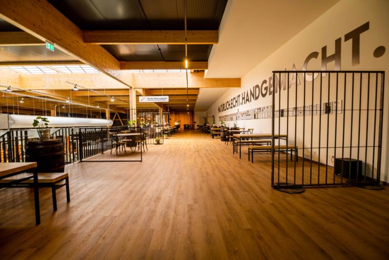 Gastronomiebereich auf der Galerieebene mit Holzboden, Tischen und sichtbaren Holzbindern, an der Wand der Schriftzug „Natürlich echt handgemacht"