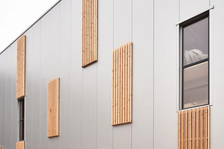 Detailaufnahme der Hallenfassade mit vertikalen Holzlamellen als gestalterische Elemente vor grauer Trapezblech-Verkleidung.