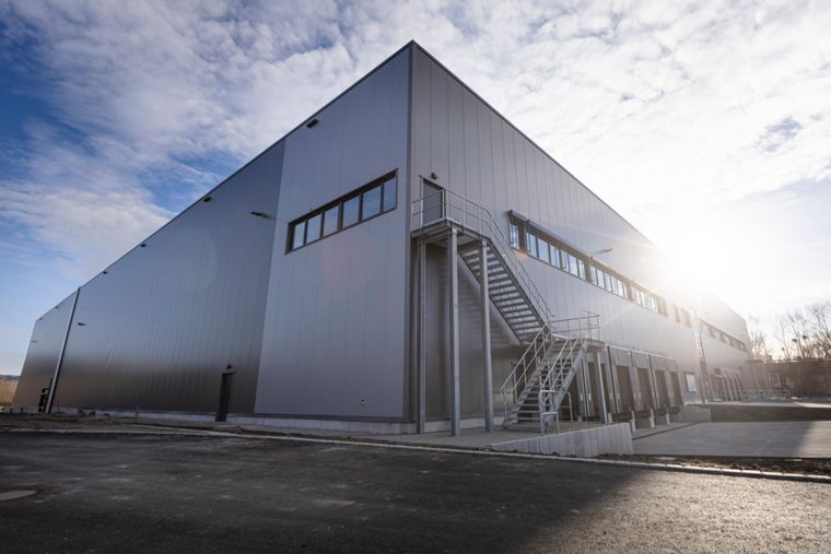 Nahaufnahme der Gebäudeecke der Logistikhalle mit außenliegender Stahltreppe im Gegenlicht und grauer Metallverkleidung.