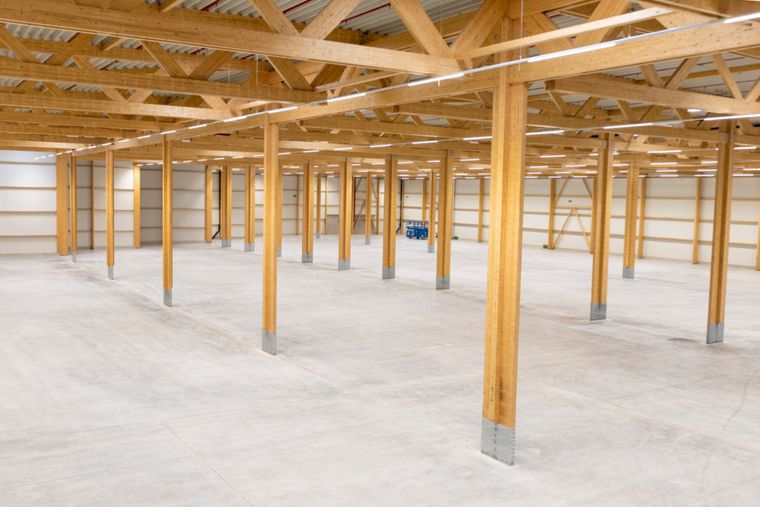 Innenansicht der Logistikhalle aus einer Ecke, mit dem gleichmäßigen Raster aus Holzstützen und Fachwerkträgern sowie dem weitläufigen, leeren Betonboden.